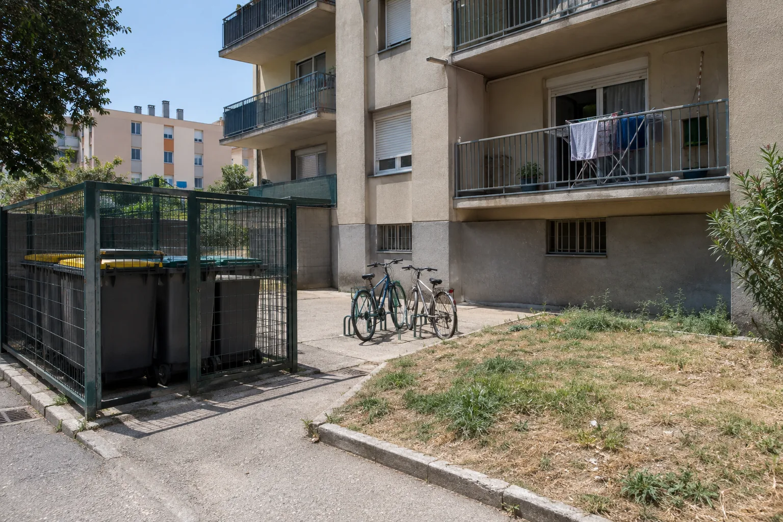 Cour d'immeuble avec local à poubelles, râtelier à vélos et pelouse fatiguée